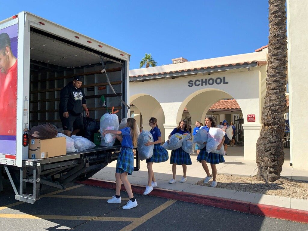 Students carry boxes from a truck outside a school building with arches and a palm tree.