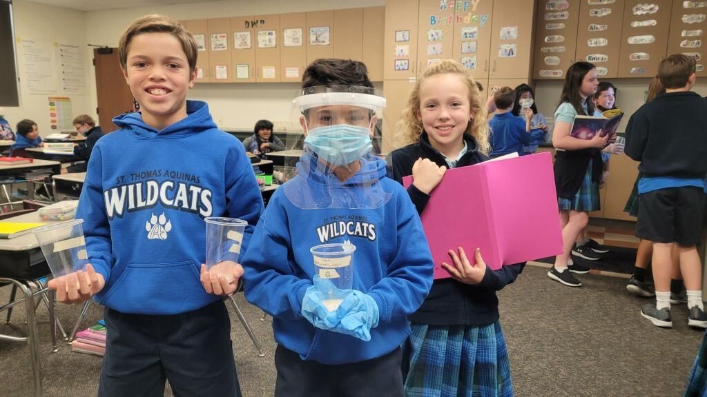Three students in blue “WILDCATS” sweatshirts hold objects in a classroom, one wearing a face shield.
