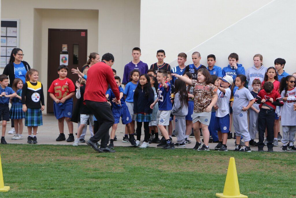 Children in blue uniforms gather outdoors while a man in red speaks to them near a school building.