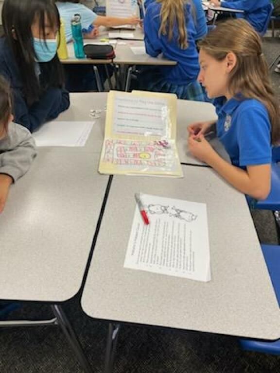 Students in blue shirts work at a classroom table with worksheets and a folder between them.