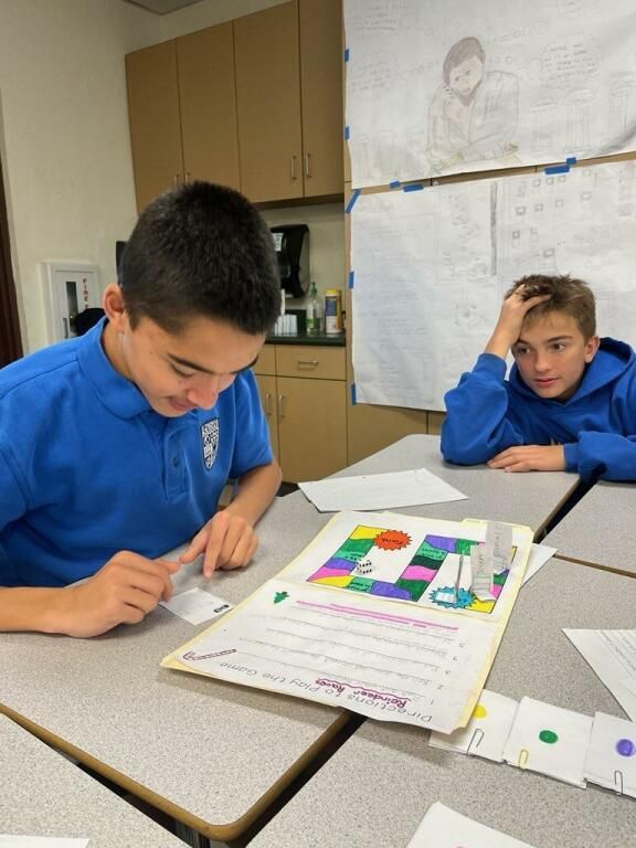 Two students in a classroom work on a colorful worksheet at a table.