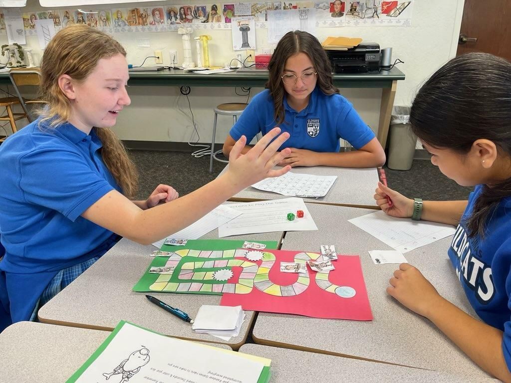 Students in blue shirts collaborate around a colorful board game at a classroom table.