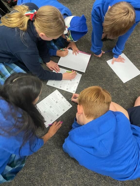 Children in blue uniforms huddle on carpet, writing on worksheets with pencils.