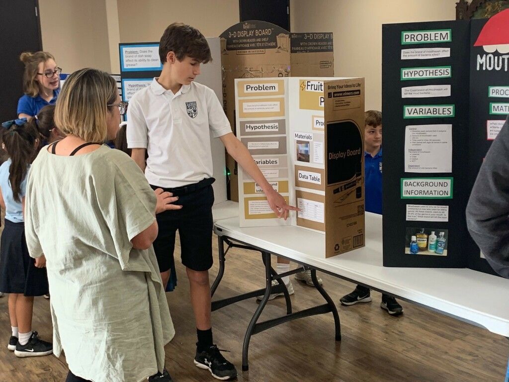 Students viewing a science fair display board with a presenter pointing at project details.