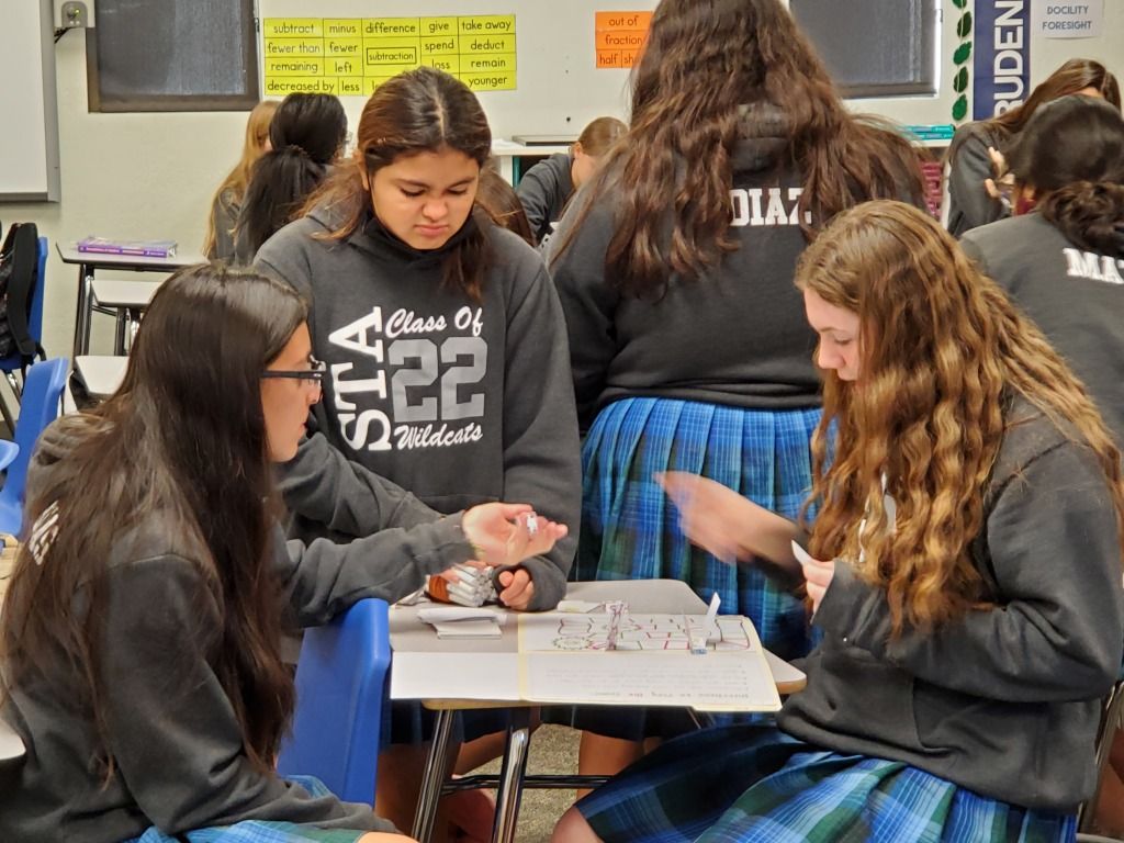 Students play a board game at a classroom table, focused on the game pieces.
