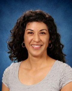 Smiling person with curly dark hair in a gray top against a blue background