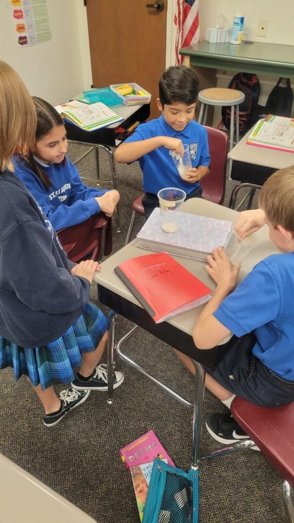Students in blue uniforms gather around a classroom table, using cups and materials for a hands-on activity.