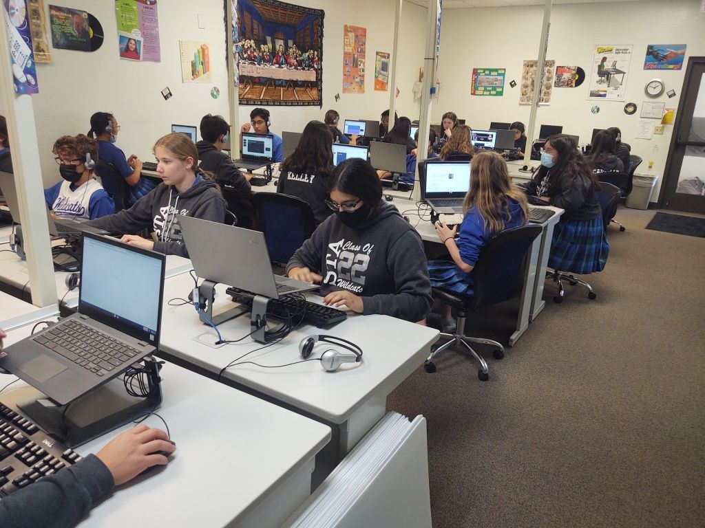 Students working on laptops in a classroom computer lab with rows of desks and wall posters.