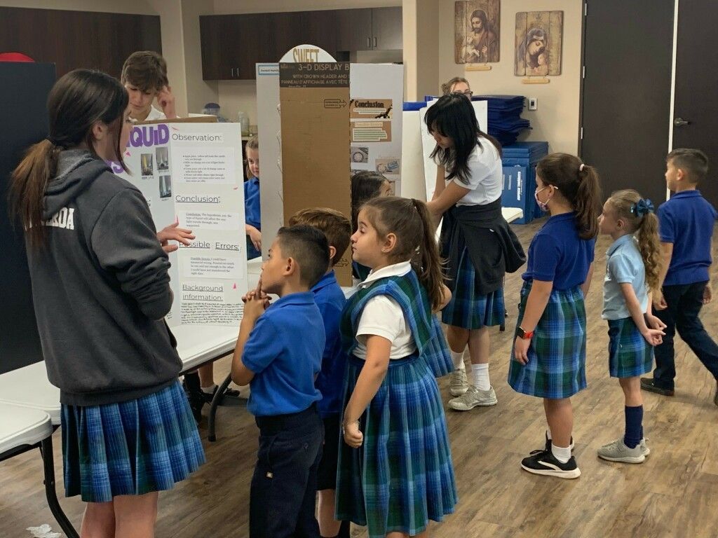 Children in blue plaid uniforms gather around a classroom poster during an activity.