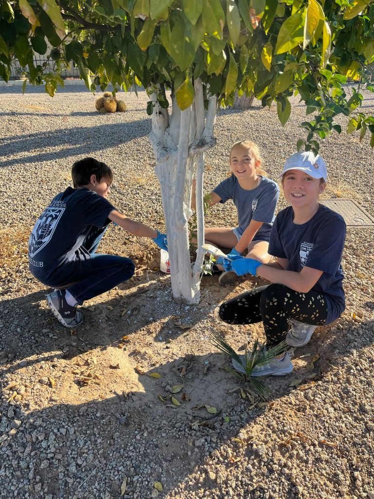 Three people crouch under a tree in a gravel orchard, posing and smiling at the camera.