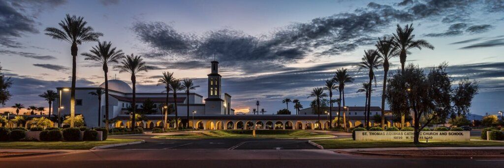 Dusk view of a mosque with minarets, palm trees, and lit fountains under a cloudy sky
