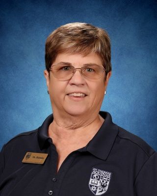 Smiling woman in glasses wearing teal jacket and name badge against a blue background