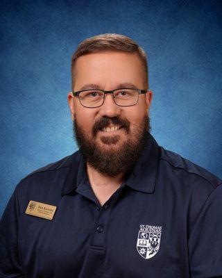 Smiling man in white shirt and striped tie with name badge against a blue studio backdrop