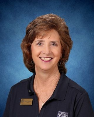 Smiling woman in a floral blouse and name badge against a blue studio backdrop