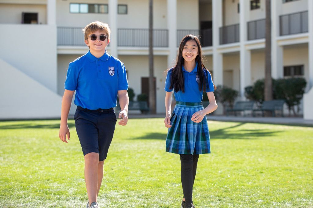 Two students in blue uniforms walking on a sunny school campus lawn