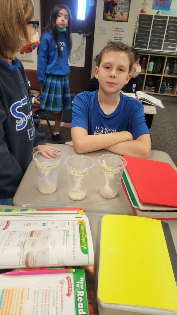 Student seated at classroom table with cups and open notebooks, another student in background