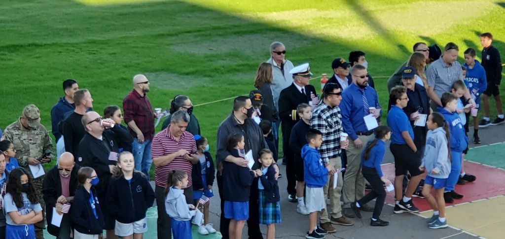 Crowd of people in casual clothes standing on a walkway beside a grassy field