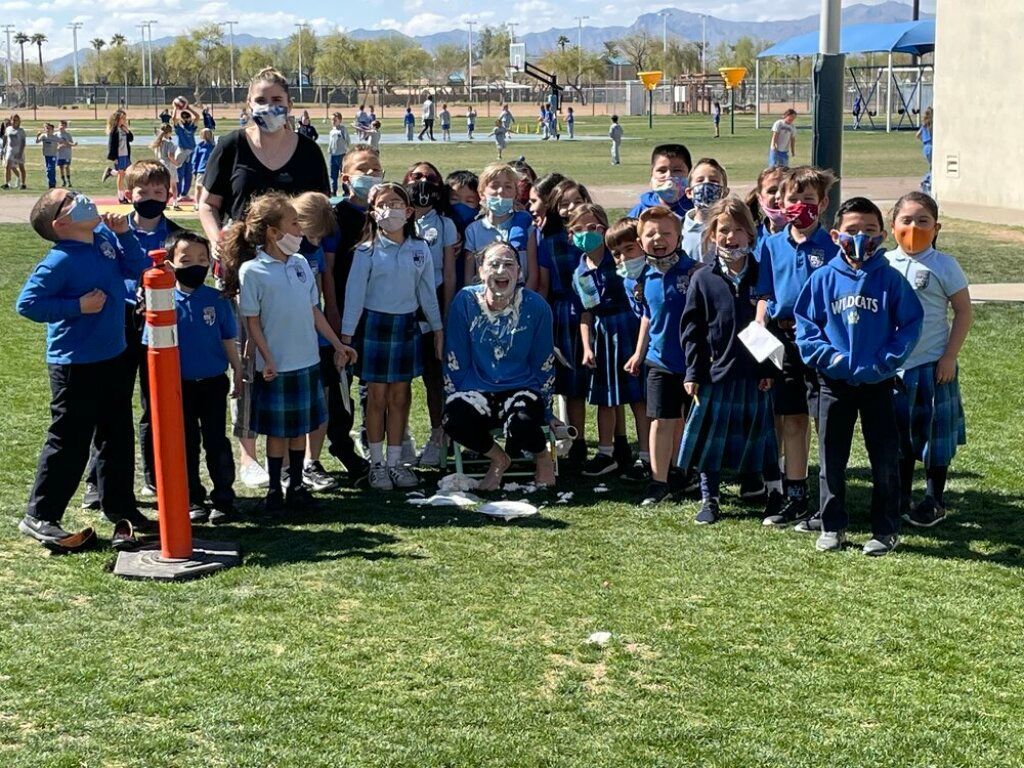 Children and adults posing on a grassy sports field, many in blue uniforms with a mountain backdrop.