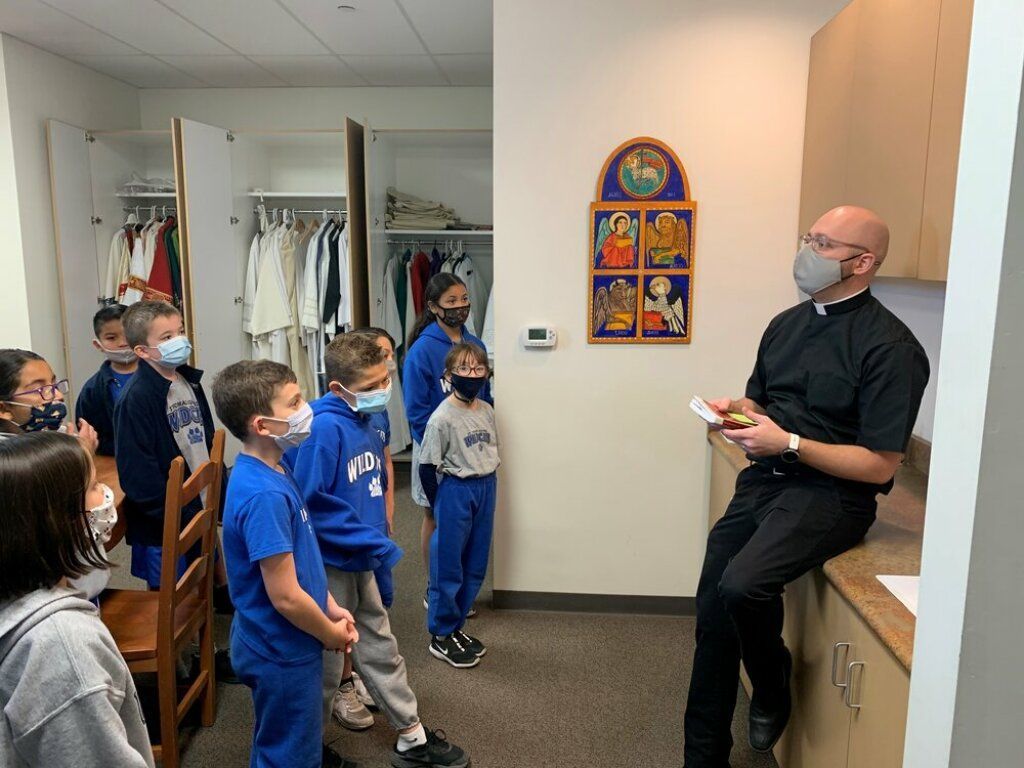 Teacher in black shirt speaking to a small group of children in a classroom hallway.