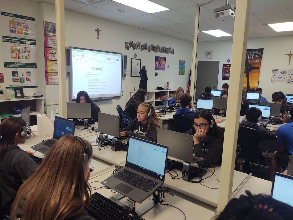 Students using laptops in a classroom during a lesson, with a projected screen at the front.