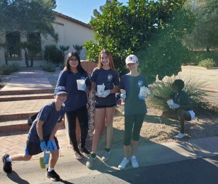 Four people outdoors by a tree, holding cups, posing on a sunny walkway.