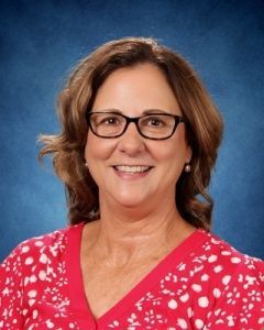 Smiling woman with glasses in a red patterned blouse against a blue studio backdrop