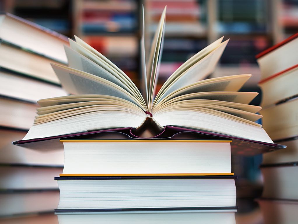 Open book atop stacked books, with pages fanned out against a blurred bookshelf background