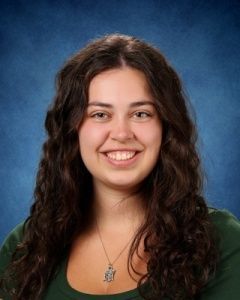 Smiling woman with long dark curly hair in a green top against a blue studio backdrop