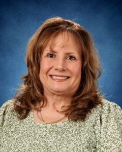 Smiling woman with shoulder-length light brown hair against a blue studio backdrop, wearing a green patterned top