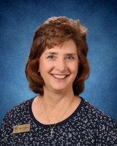 Smiling woman in a floral blouse and name badge against a blue studio backdrop