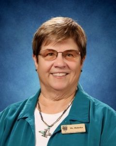 Smiling woman in glasses wearing teal jacket and name badge against a blue background