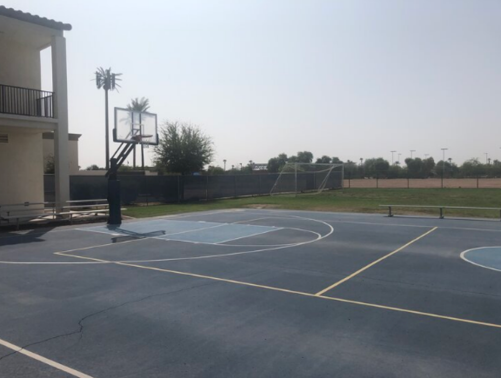 Empty outdoor basketball court beside a building, with hoops and painted court lines under a bright sky