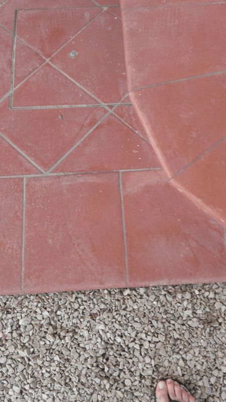 Pavimento in piastrelle rosse con disegno geometrico accanto a ghiaia grigia; è visibile un piede nudo.