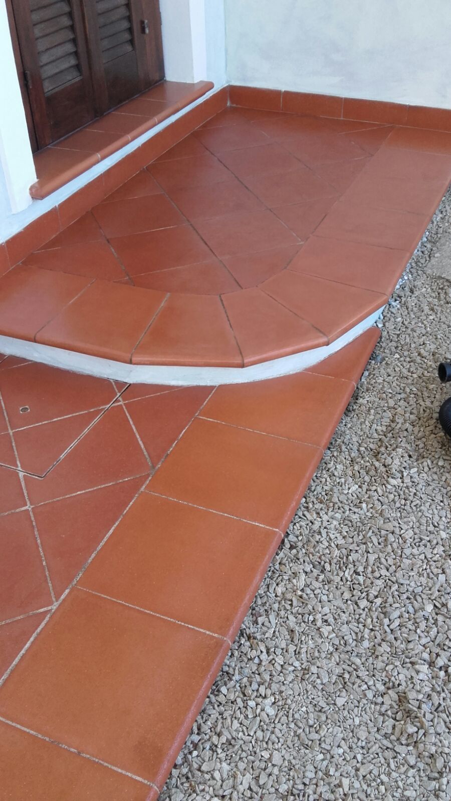 Patio piastrellato in terracotta con bordo curvo, adiacente alla ghiaia, vicino a un edificio.