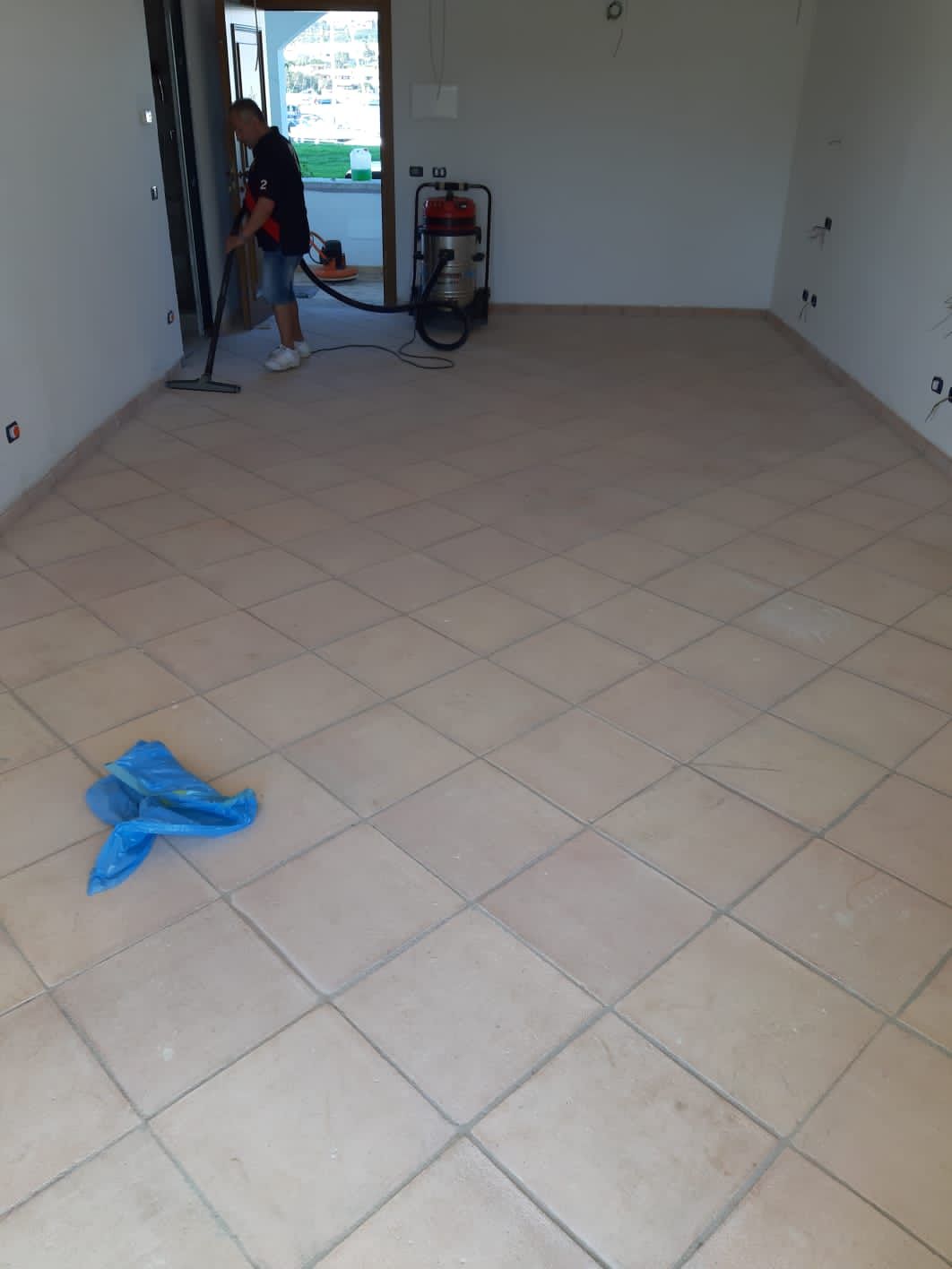 Persona che passa l'aspirapolvere in una stanza con pavimento in piastrelle beige, copertura protettiva blu sul pavimento e porta sullo sfondo.