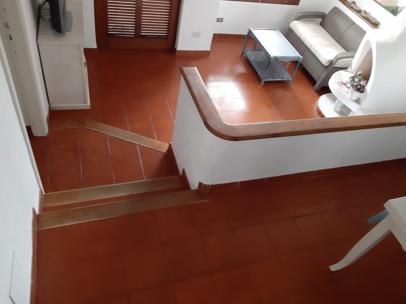 Guardando in basso verso le scale con piastrelle in terracotta e gradini in legno che conducono a una zona soggiorno con pareti bianche e un divano.