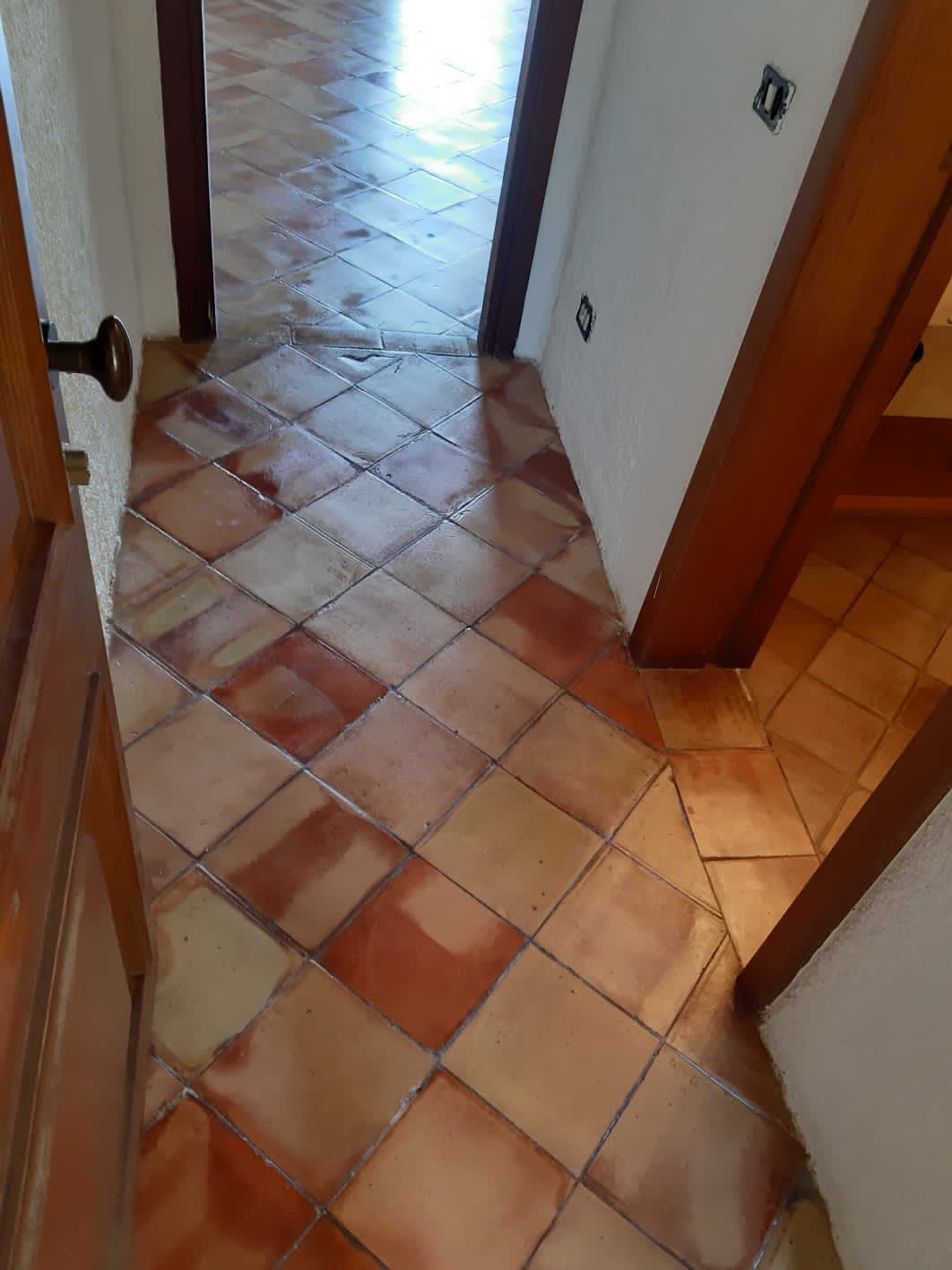 Corridoio con piastrelle quadrate rosso-marroni posate in diagonale. Porte con stipiti in legno e pareti bianche.