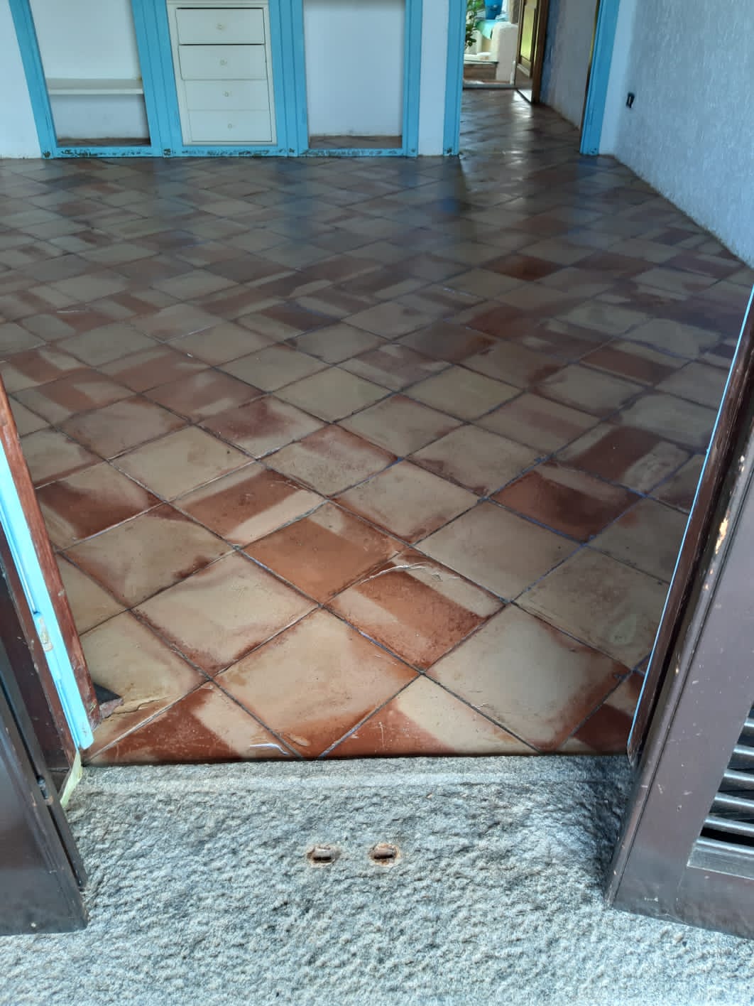 Pavimento piastrellato marrone e beige, visto dall'ingresso con porte marroni chiuse. Sullo sfondo, cornici delle porte turchesi.