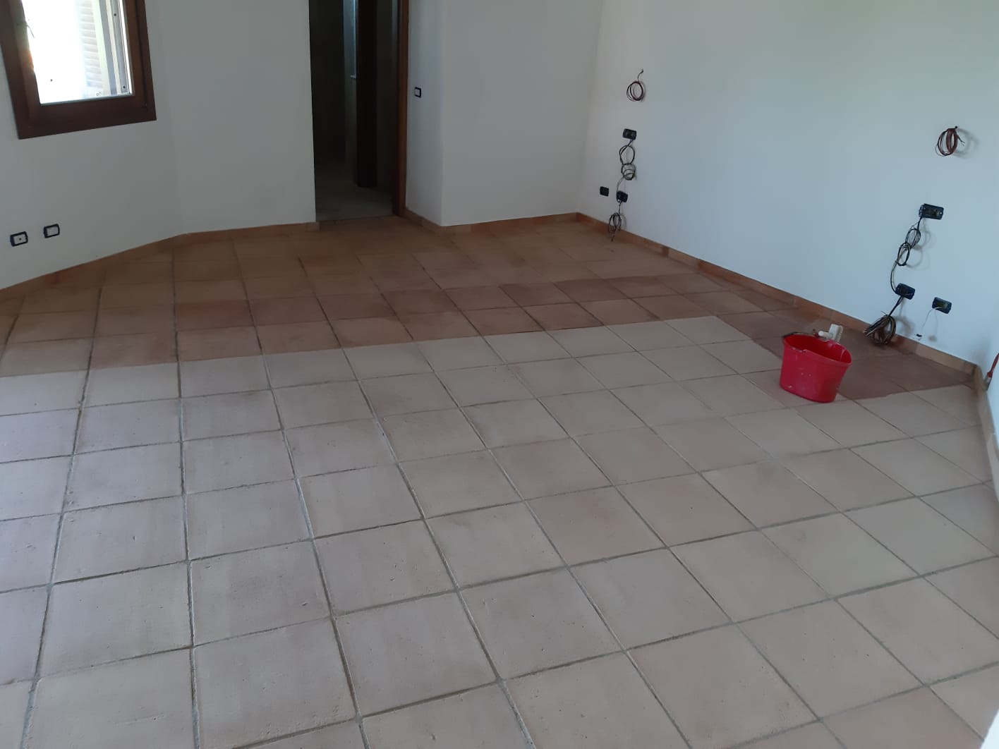 Stanza con pavimento in piastrelle beige e pareti bianche. Un secchio rosso è appoggiato sul pavimento.