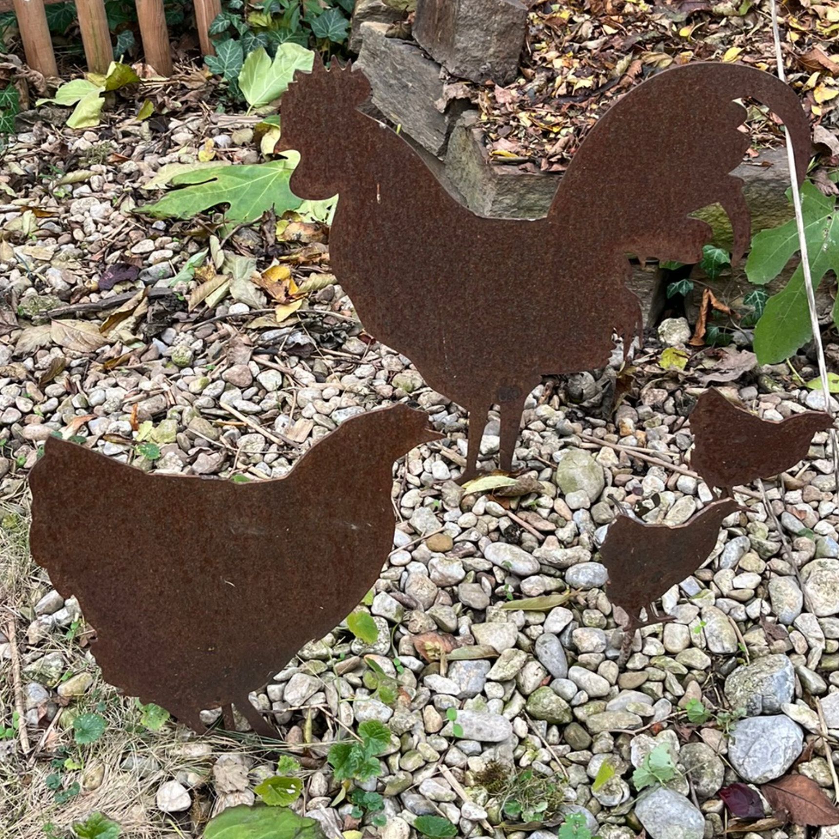 Rostige Metallfiguren von Hahn, Henne und Küken als Gartendekoration auf Kies.