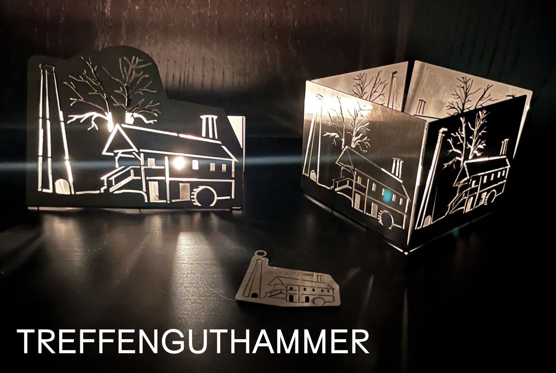 Zwei metallene Kerzenhalter mit ausgeschnittenen Haus- und Baummotiven, sanft beleuchtet.