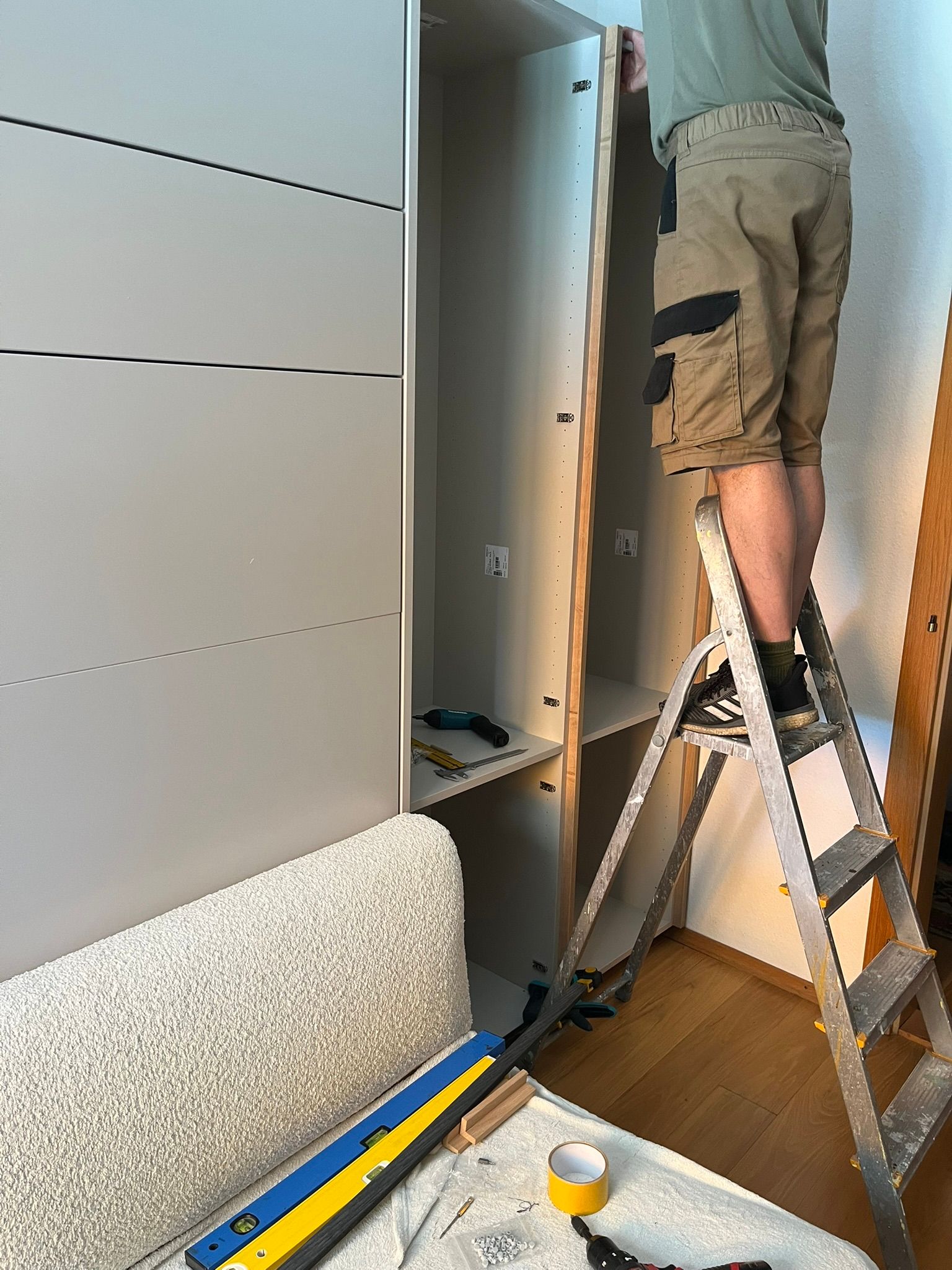 Person auf einer Trittleiter montiert einen hohen, hellen Schrank; Innenaufnahme.