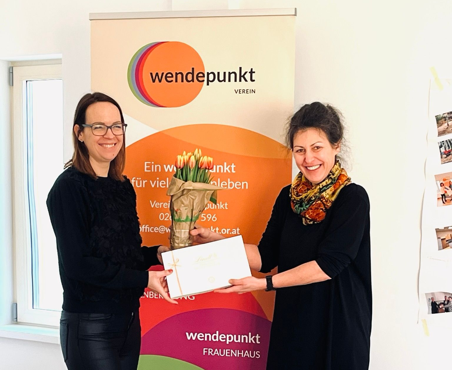 Zwei Frauen tauschen vor einem Banner mit der Aufschrift „Wendepunkt“ Blumen und ein Geschenk aus.