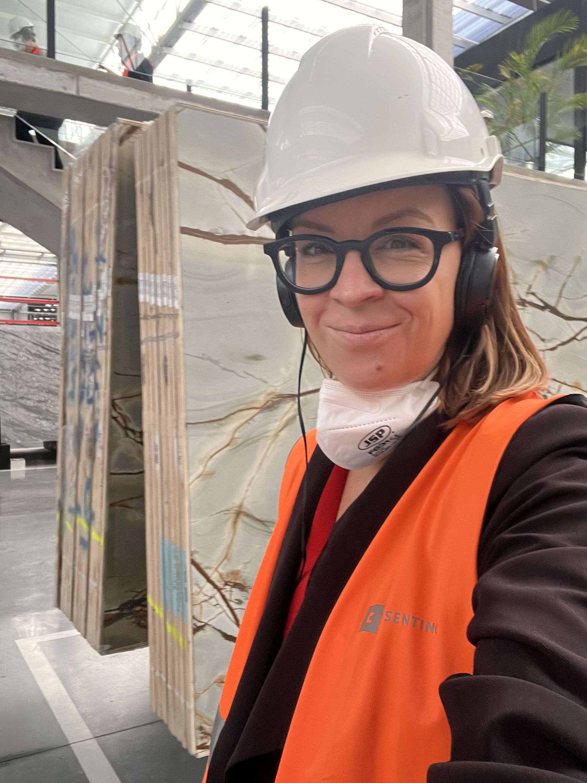 Eine Frau mit Schutzhelm, Sicherheitsweste und Brille lächelt in einer Fabrikhalle;