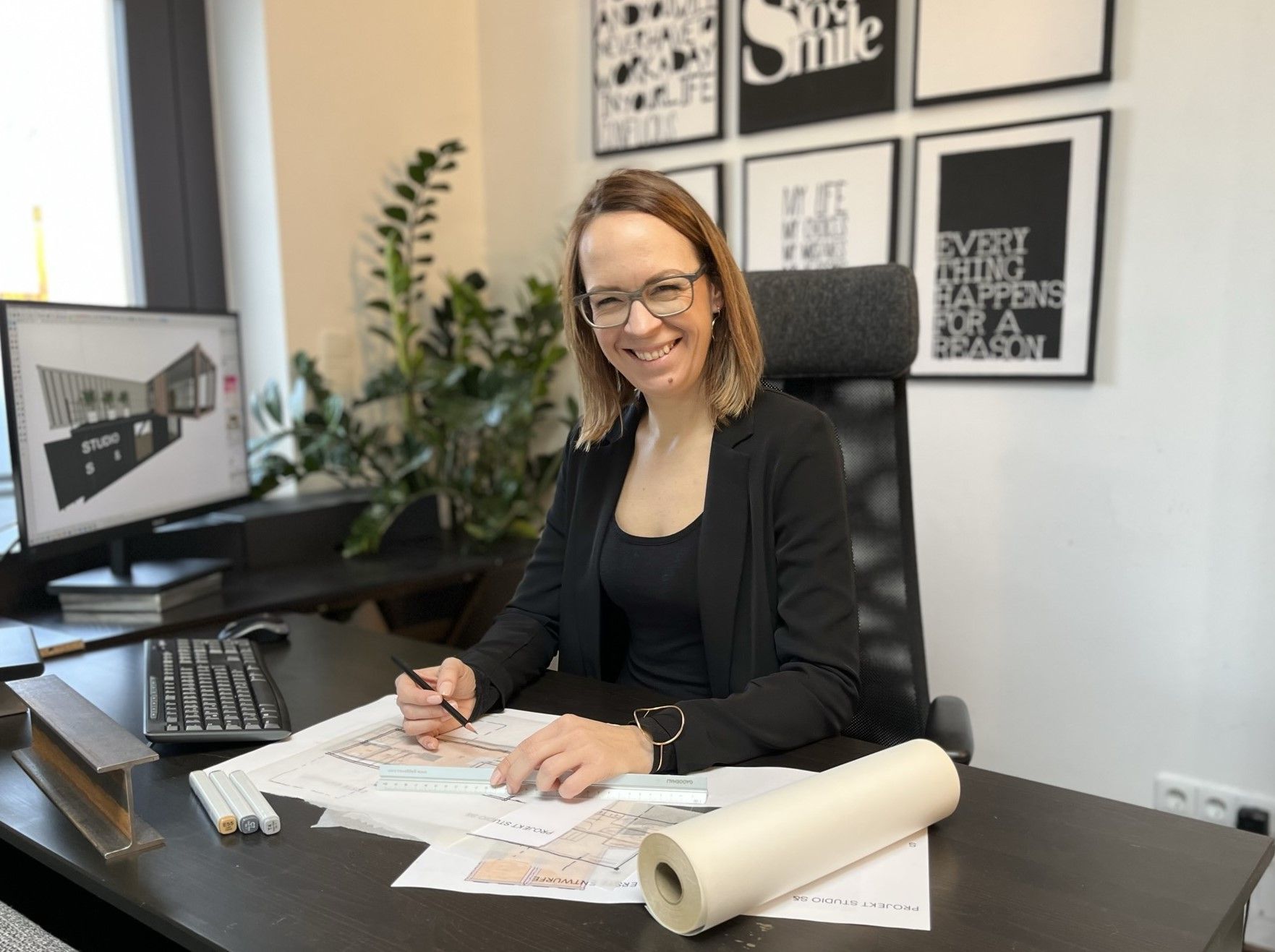 Eine Frau mit Brille sitzt an einem Schreibtisch mit Bauplänen, Computer und Architekturentwürfen.
