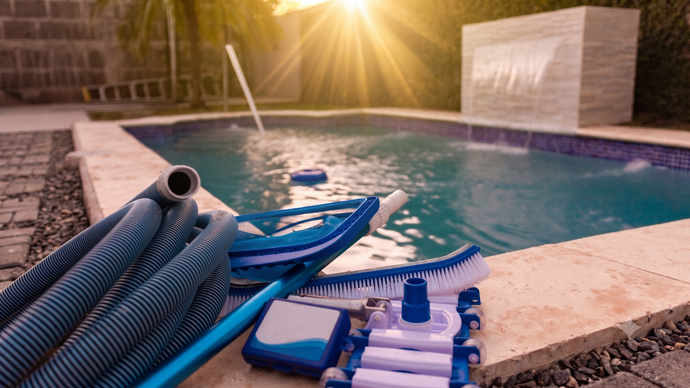 Pool-cleaning supplies beside a sunlit backyard pool, with hoses and brushes on the deck