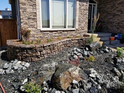 A stone wall is surrounded by rocks and plants in front of a house.
