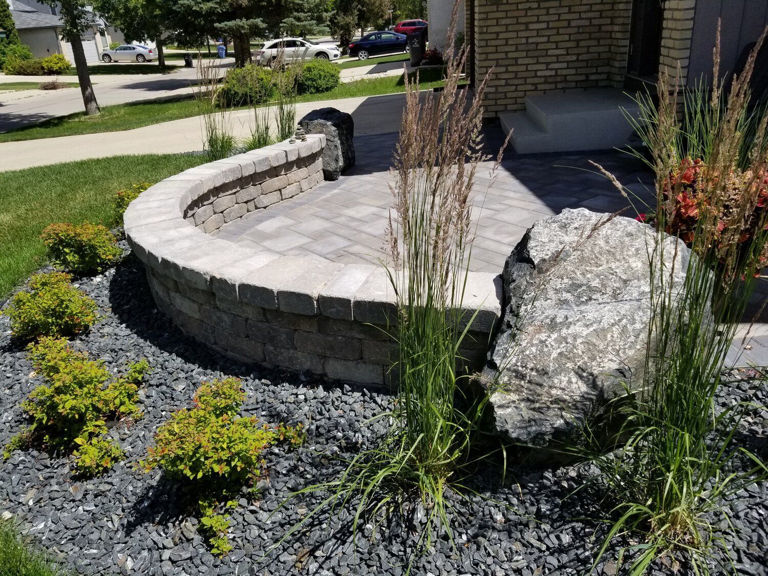 A circular stone wall is surrounded by gravel and plants in front of a house.