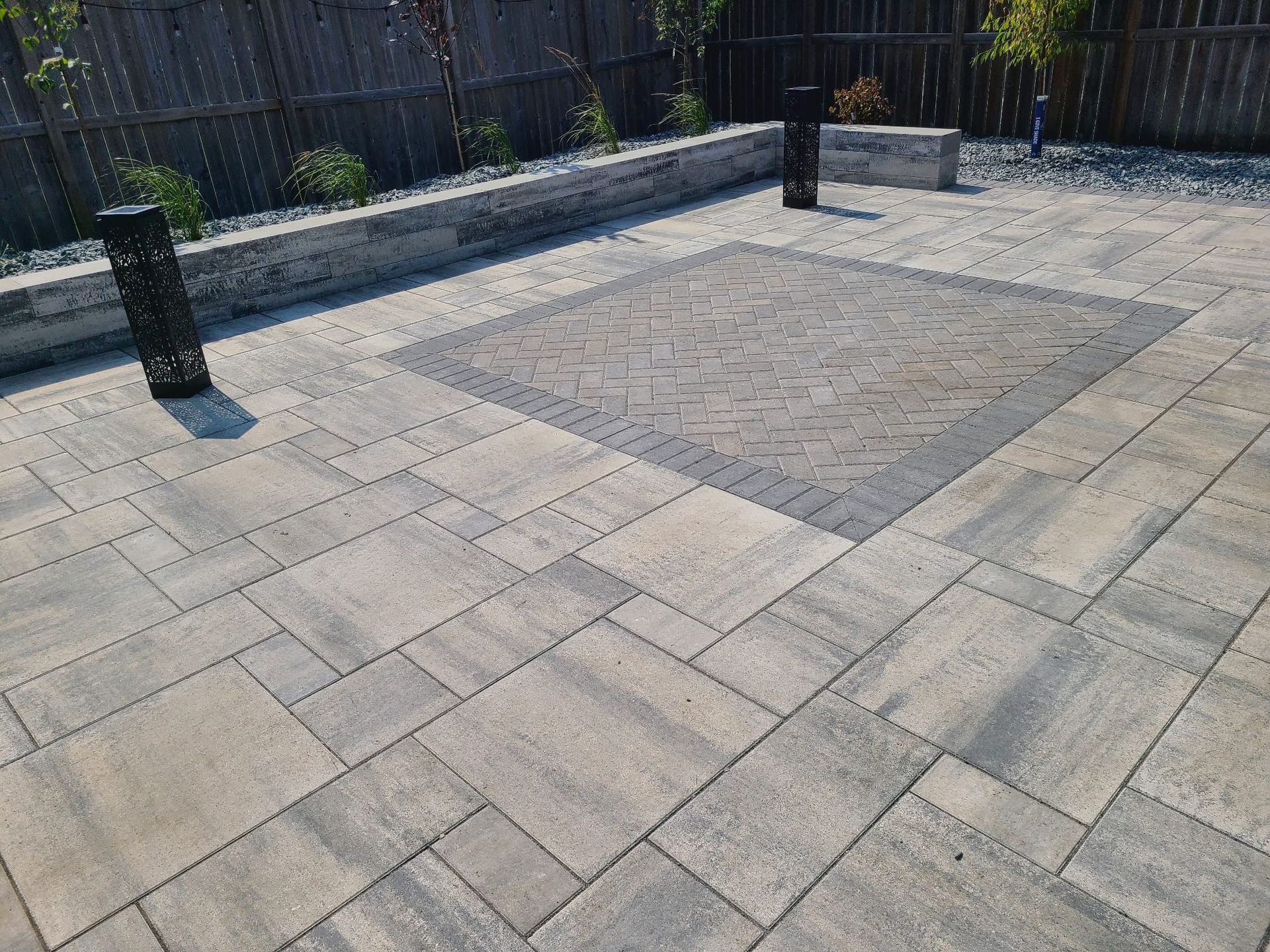 A paver patio with a fence in the background and a square in the middle.