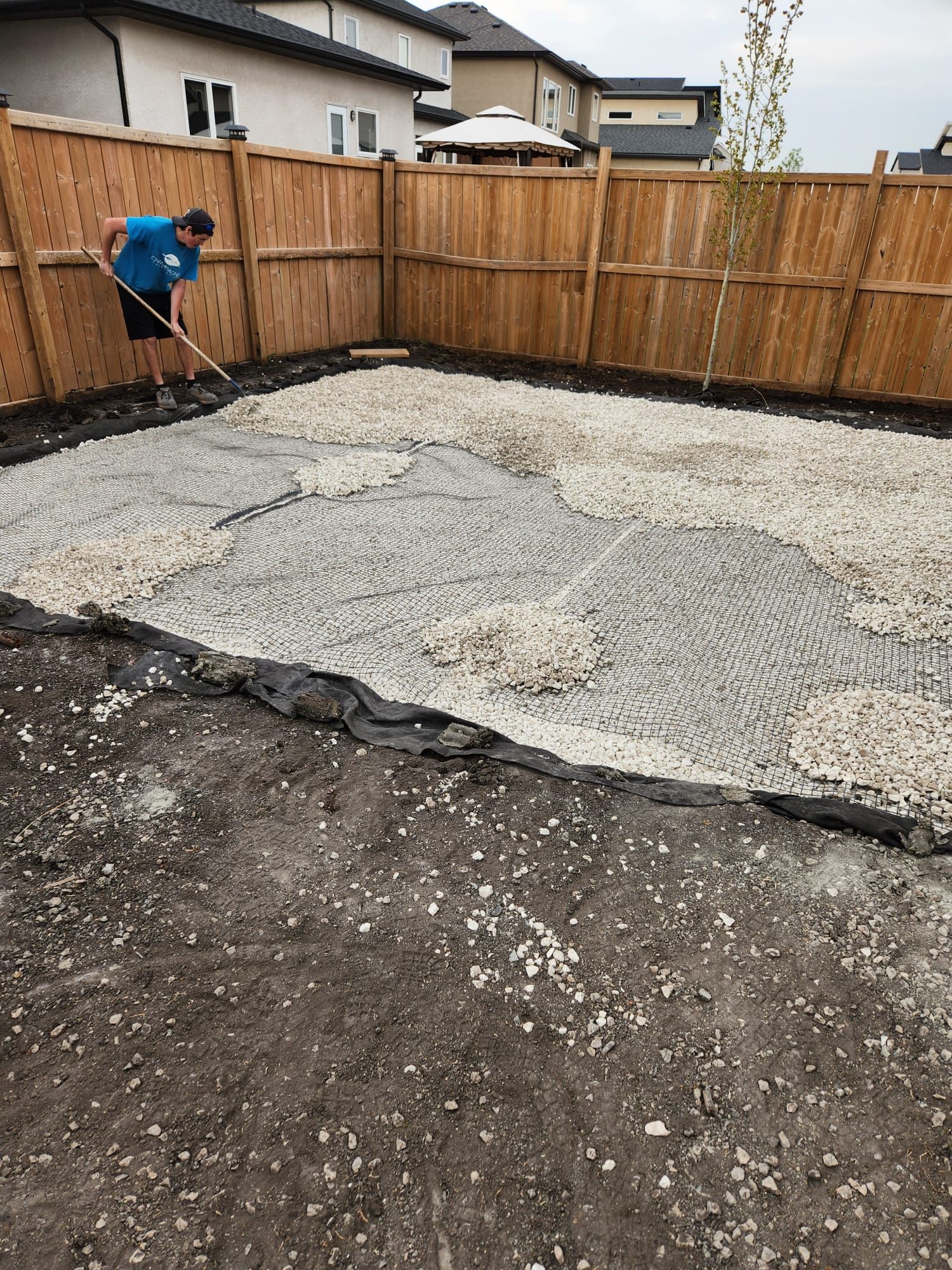 A man is raking dirt in a backyard next to a wooden fence.